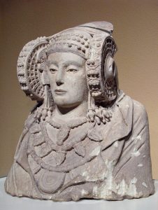 Foto de la Dama de Elche, un busto esculpido en piedra que retrata una señora que lleva en la cabeza un tocado grande e intrincado además de varios collares.