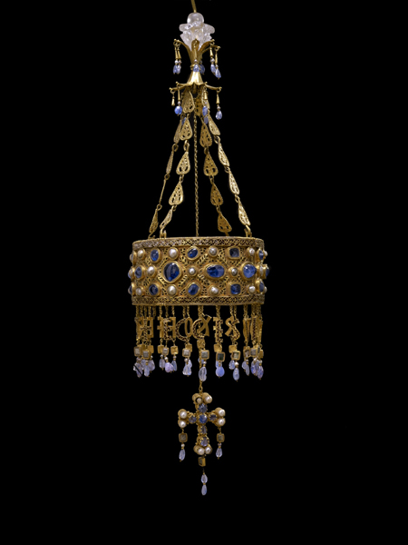 Foto de un objeto de orfebrería hecho de oro y piedras preciosas blancas rojas y azules; una corona circular está suspendida por cadenas decorativas de un capitel en cristal, de la corona misma penden otras cadenas con letras formadas en oro que representan la dedicación votiva, y colgando en el centro está una cruz decorada con perlas y zafiros.