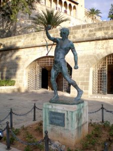 foto de una estatua en bronce de un hondero balear; tiene su hondo en el brazo derecho como si acabara de lanzar un proyectil.