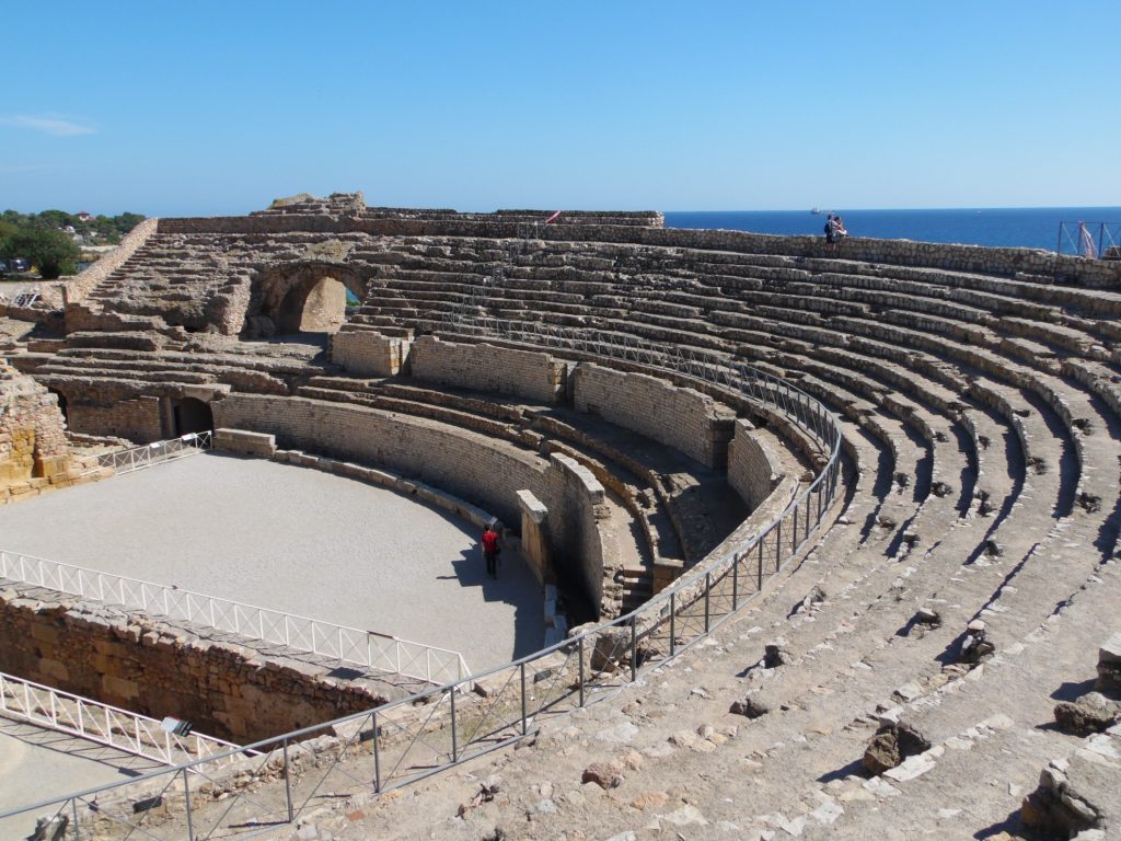 foto del anfiteatro romano en Tarragona donde se ve los grados de asientos hechos de piedra y cemento en semicírculo sobre el espacio de arena donde luchaban los gladiadores; el mar Mediterráneo es visible detrás.