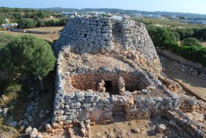 foto de talayot en Menorca, mostrando un gran edificio de piedra; la gran sala de entrada tiene dos columnas que apoyarían al techo, y hay una ancha torre circular detrás