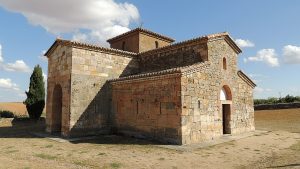 Exterior de la iglesia visigoda, mostrando un arco en forma de herradura sobre una puerta, el hueco para una ventana semicircular sobre otra puerta, y otras ventanas muy estrechas en las paredes de piedra.