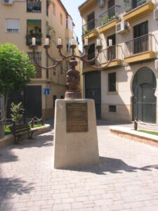 foto de un menorá grande con brazos curvados de metal, sobre un pedestal rectangular de cemento, en medio de una pequeña plaza en la zona histórica de la ciudad
