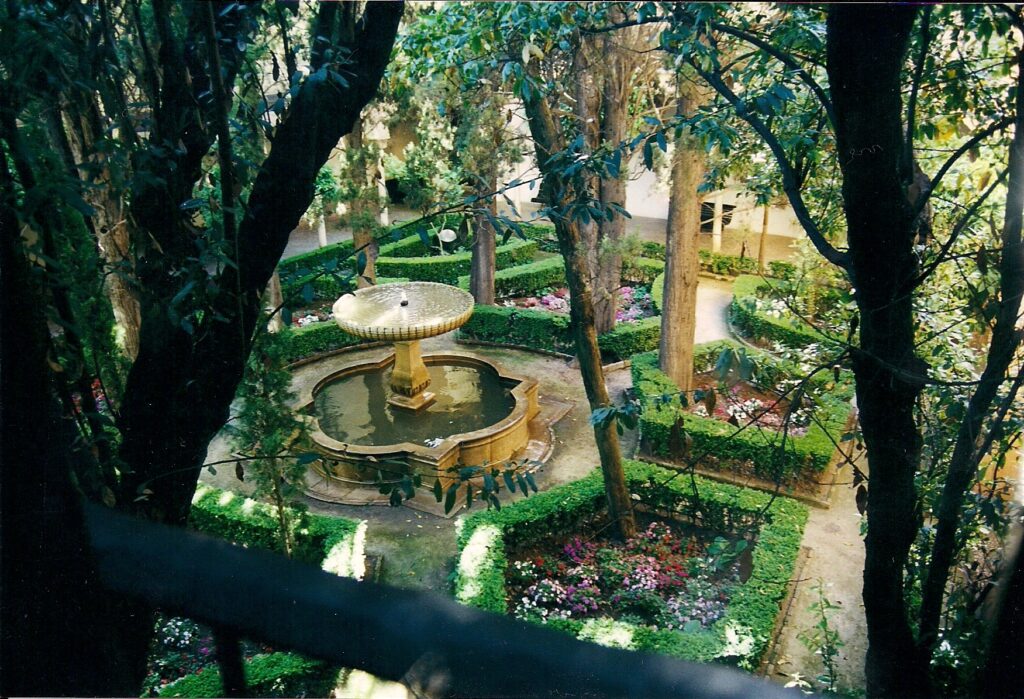 Foto desde arriba mostrando un patio interior con una fuente en el centro y jardines geométricos con flores, arbustos y naranjos, árboles que dan una sombra refrescante.