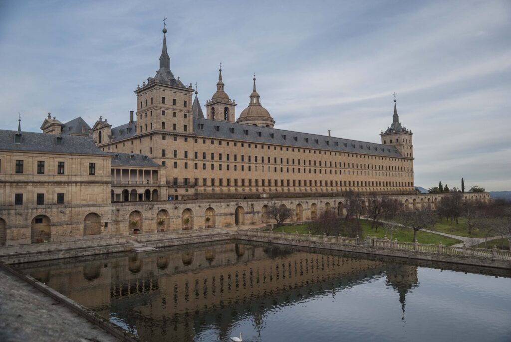 foto del exterior de El Escorial, con su vasta fachada de ventanas rectangulares, frente a una alberca ("pool") rectangular y parte del jardín