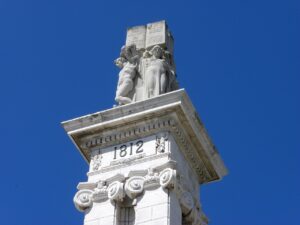 foto del punto más alto del monumento, que muestra la fecha 1812 sobre unas columnas iónicas, y encima dos figuras neoclásicas levantan un libro con texto de la Constitución