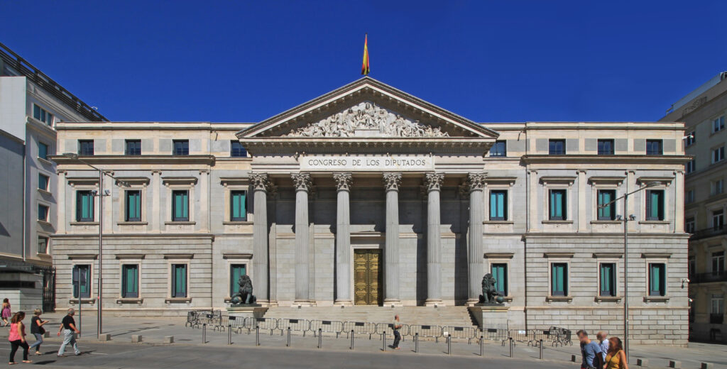 foto de un edificio neoclásico de tres pisos con ventanas rectangulares y seis pilares de dos pisos en altura delante de su puerta de entrada; encima de la puerta dice Congreso de los Diputados
