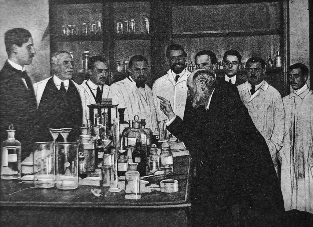 foto de nueve hombres detrás de una mesa llena de frascos y microscopios, escuchando al profesor Ramón y Cajal, que está a la derecha