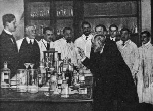 foto de nueve hombres detrás de una mesa llena de frascos y microscopios, escuchando al profesor Ramón y Cajal, que está a la derecha