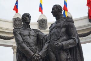 Estatua en bronce de dos hombres en uniforme militar del siglo 19 estrechándose la mano; detrás hay banderas de los países sudamericanos que ellos liberaron