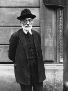 foto de un hombre con barba y bigotes blancos, llevando un traje y sombrero negros al estilo del comienzo del siglo 20