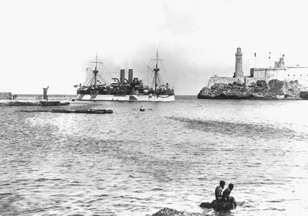 Un buque militar con dos chimineas, dos mástiles y varios cañones pasa frente al faro y murallas defensivas