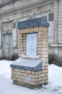 una placa de azulejos blancos y azules en un pilar de ladrillos de color claro, con letra en el idioma letón (Lithuanian) y una parte en castellano que dice "Ángel Ganivet, Granada recuerda su memoria"